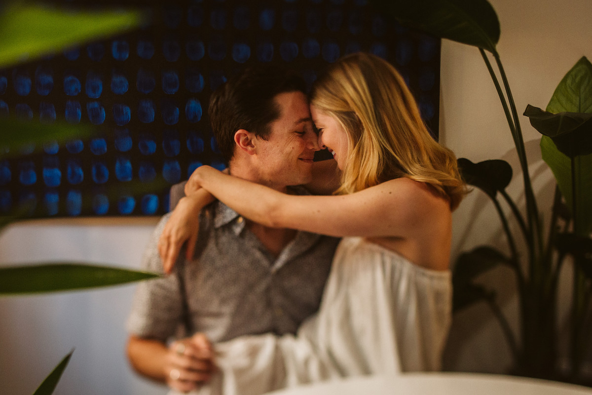 Woman sits on man's lap in front of a large black and blue painting as they cuddle. They're surrounded by large plants.