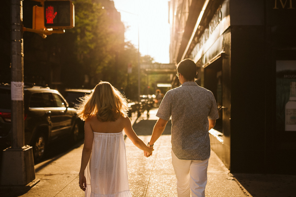 Woman in flowing, white sundress holds hands with man in blue floral shirt and white trousers as they walk down the sidewalk