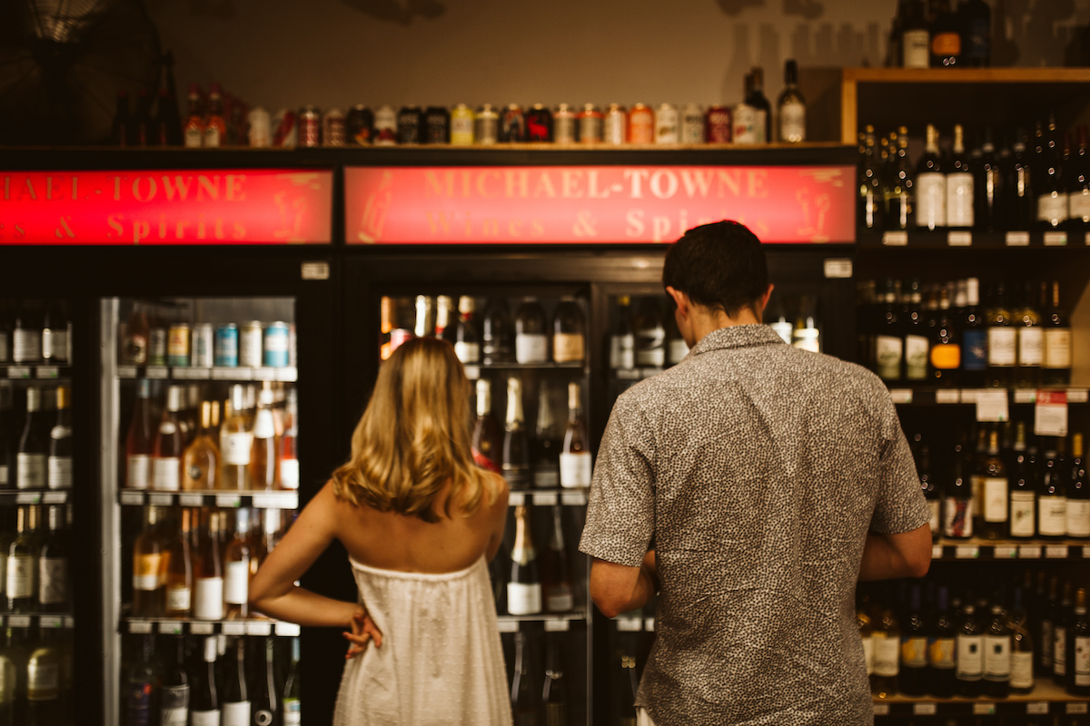 Man and woman stand in front of glass door refrigerators with various types of wine and champagne