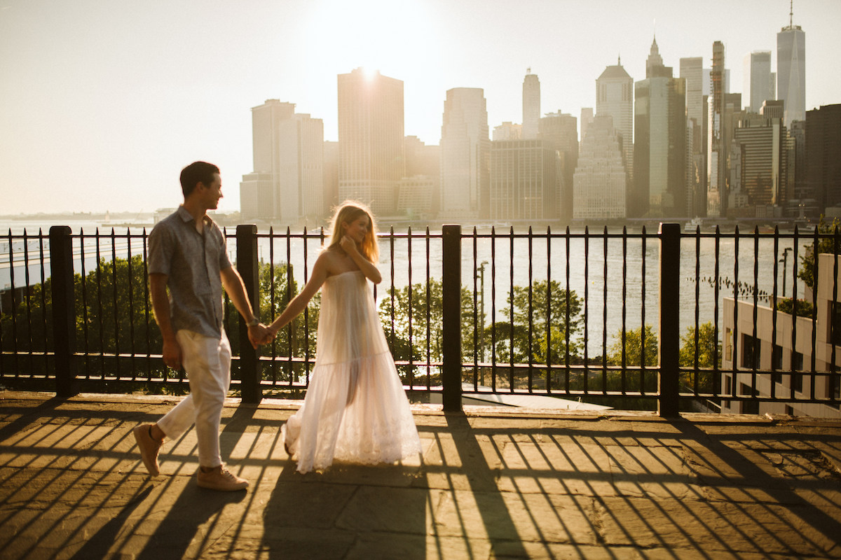 Man and woman hold hands and walk in front of a black, metal fence along waterfront. New York City skyline is behind them.