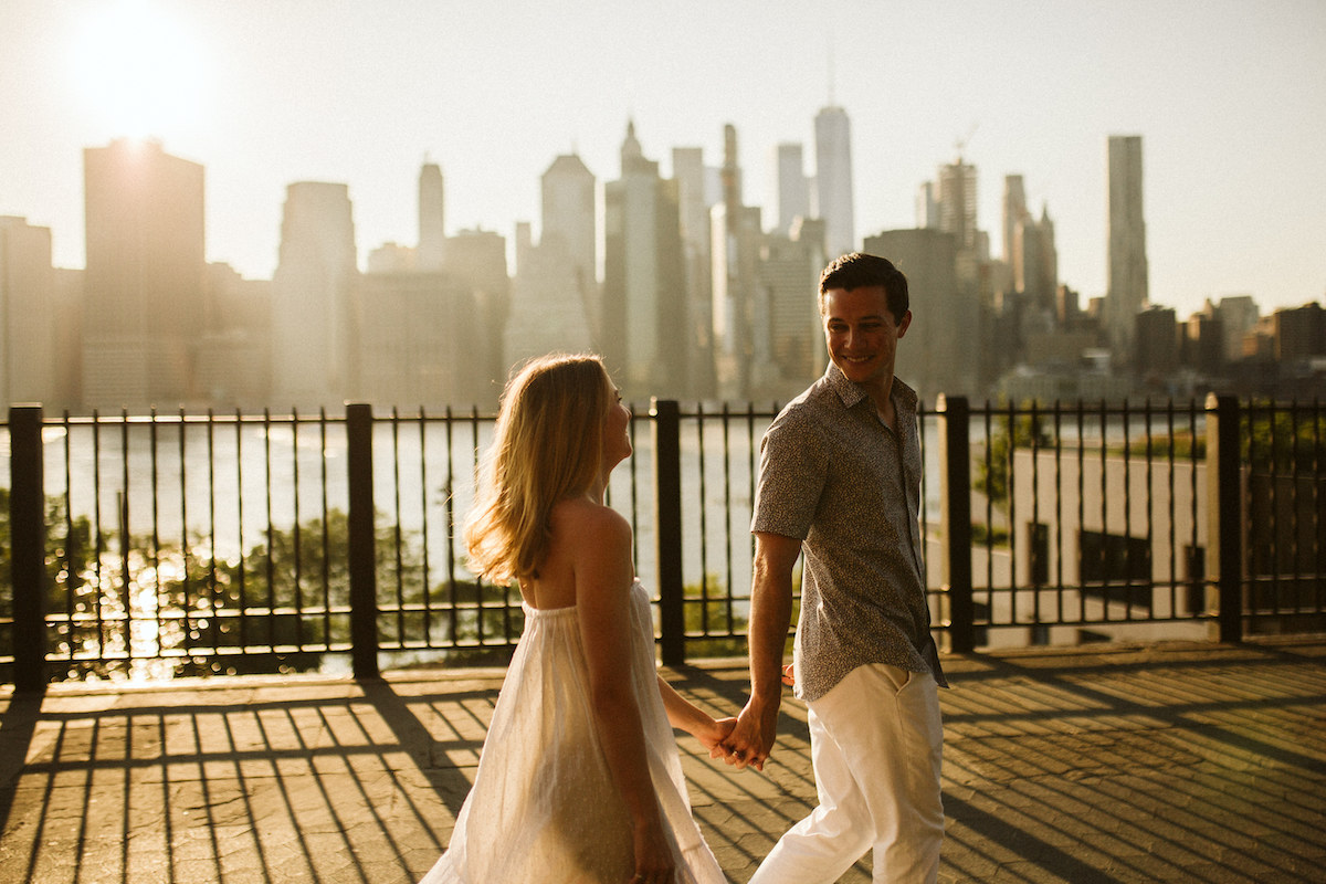 Man and woman hold hands and walk in front of a black, metal fence along waterfront. New York City skyline is behind them.