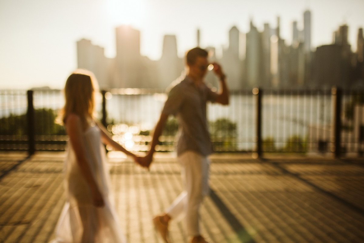 Man and woman hold hands and walk in front of a black, metal fence along waterfront. New York City skyline is behind them.