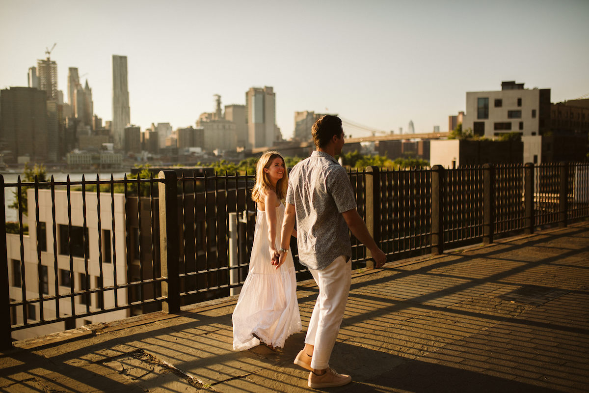 Man and woman hold hands and walk in front of a black, metal fence along waterfront. New York City skyline is behind them.