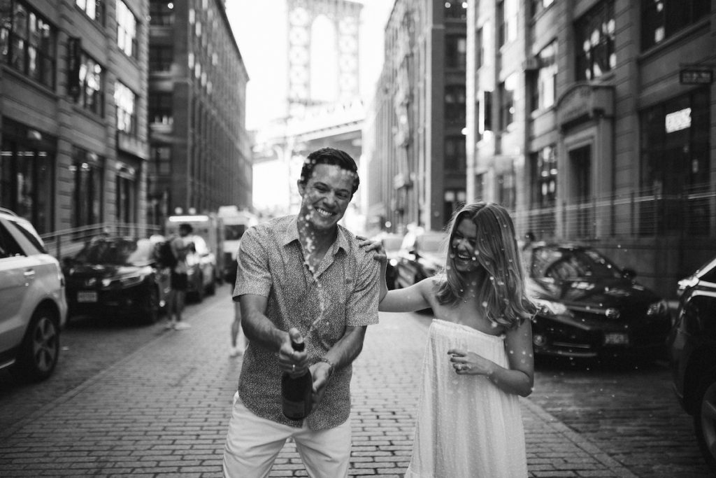 Man sprays champagne on a cobblestone street of Dumbo Brooklyn while woman places her hand on his shoulder, laughing.