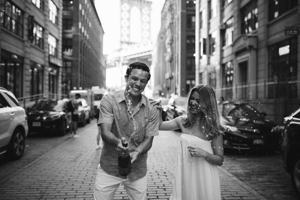 Man sprays champagne on a cobblestone street of Dumbo Brooklyn while woman places her hand on his shoulder, laughing.