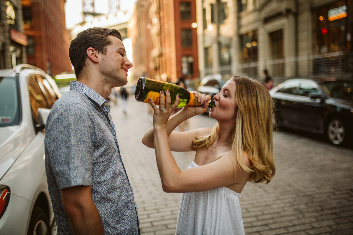 Woman in strapless white sundress drinks champagne from the bottle while man watches during Dumbo Brooklyn engagement session