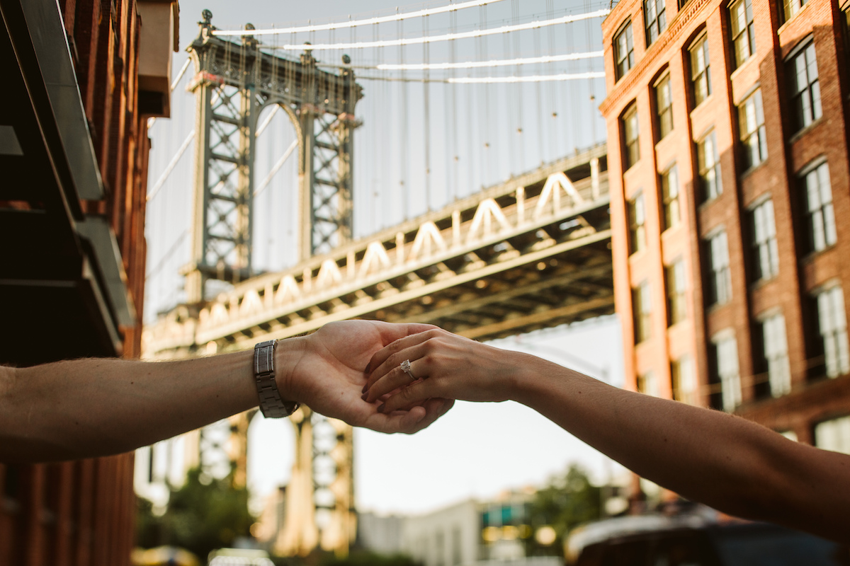 Man's arm with silver watch and woman's arm with simple, large diamond ring. Brooklyn Bridge in the background.