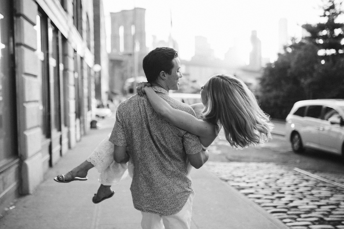 Man carries woman on the sidewalk during their engagement session in Dumbo Brooklyn near the Brooklyn Bridge