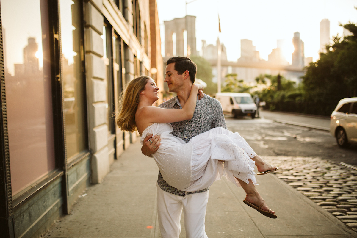 Man carries woman on the sidewalk during their engagement session in Dumbo Brooklyn near the Brooklyn Bridge