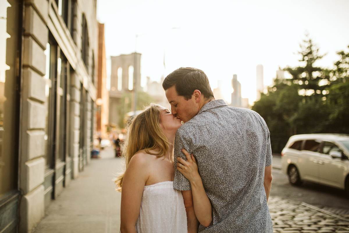 Man and woman kiss on the sidewalk during their engagement session in Dumbo Brooklyn near the Brooklyn Bridge