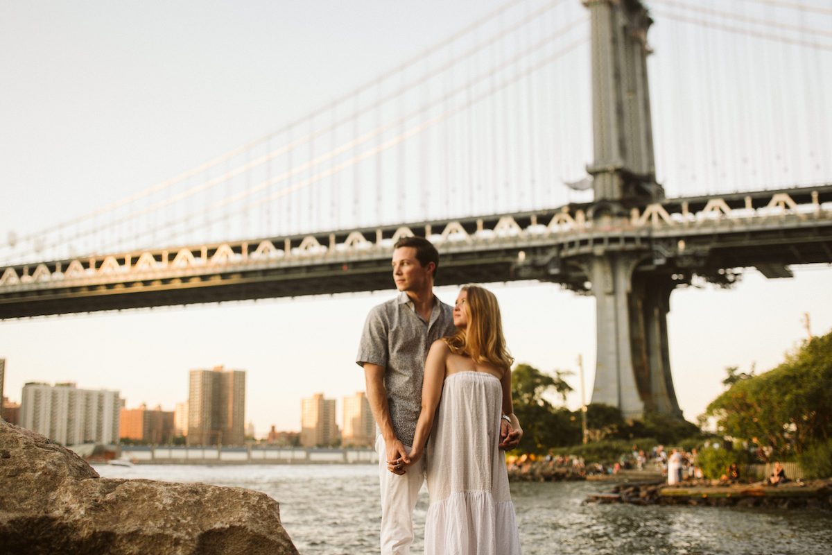 Man and woman stand gazing at the sunset under the Brooklyn Bridge during Dumbo Brooklyn engagement session