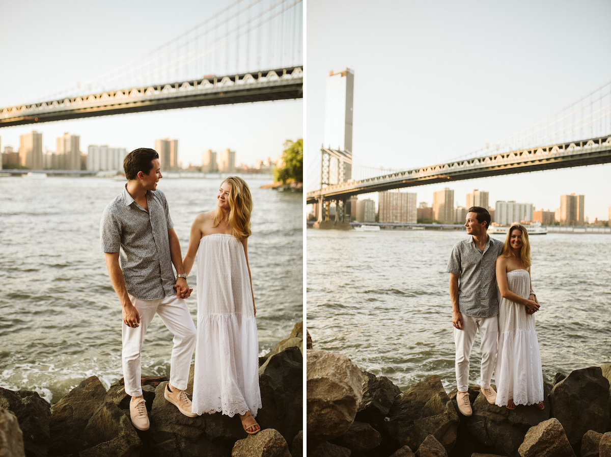 Man and woman stand on large rocks next to the East River under the Brooklyn Bridge during Dumbo Brooklyn engagement session