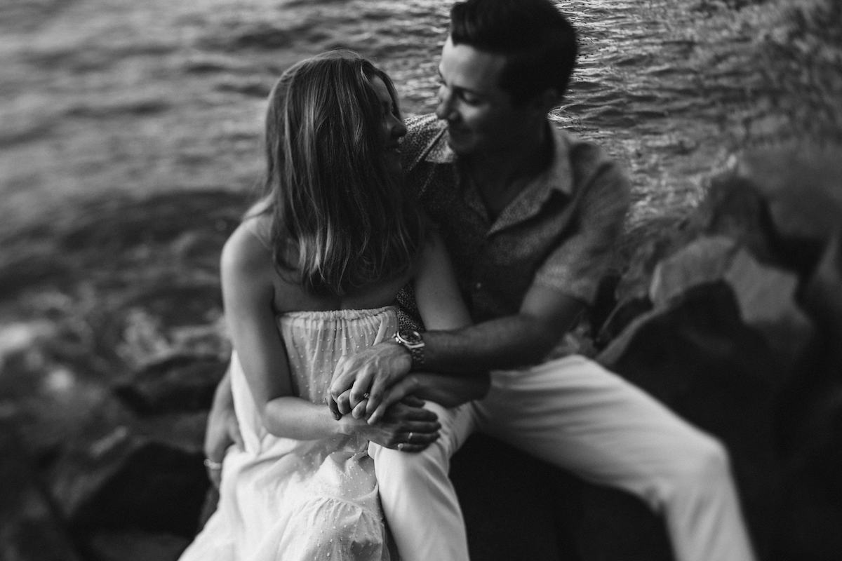 Man and woman cuddle on large rocks beside the East River in Brooklyn New York City