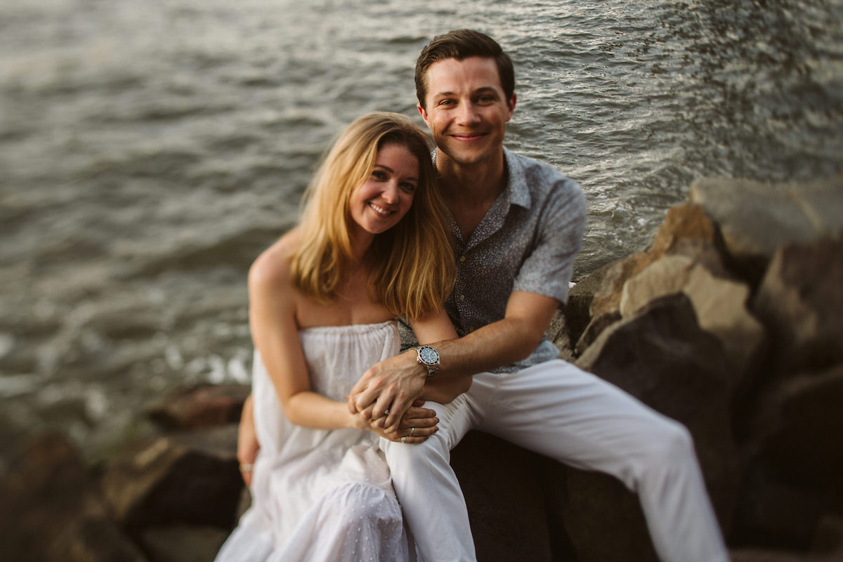 Man and woman cuddle on large rocks beside the East River in Brooklyn New York City