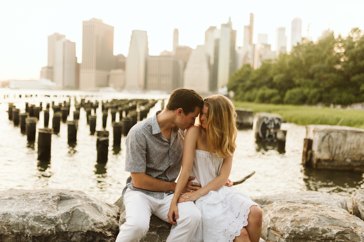 Man kisses woman's shoulder while they sit on large rocks waterside with New York City scape in background