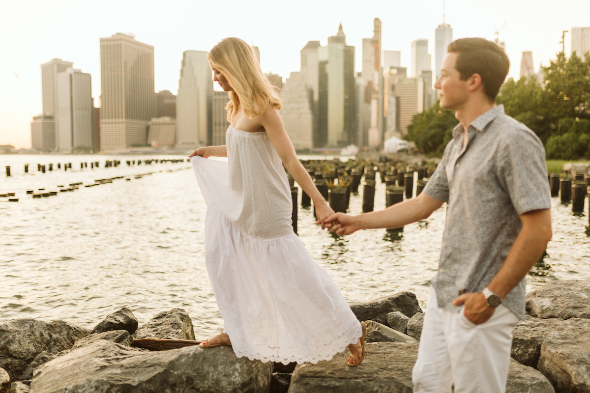 Man and woman hold hands and walk along large rocks waterside with New York City scape in background