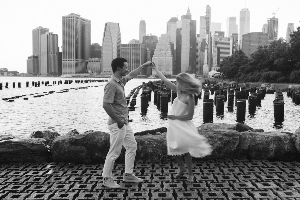 Man twirls woman in white sundress near water with New York City scape behind them