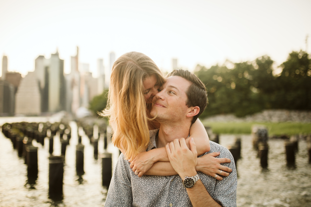 Woman cuddles man from behind during golden hour in front of New York City scape