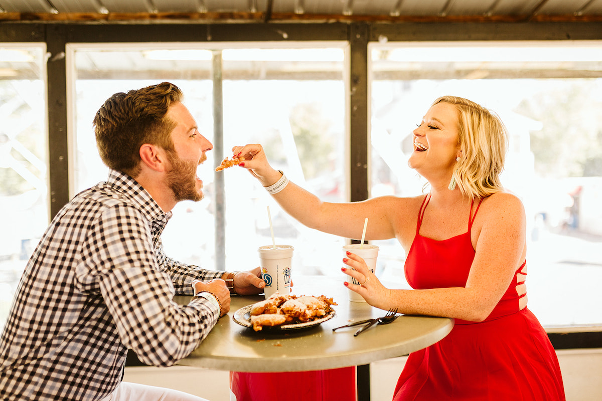 Woman reaches across a table to offer a bite of funnel cake to man