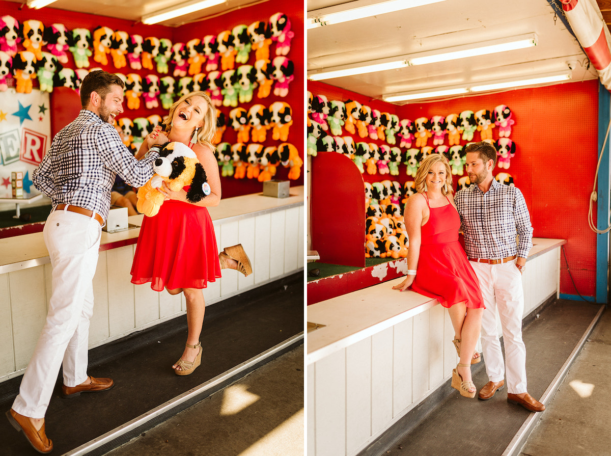 Man stands beside woman sitting on counter of carnival game with a wall of stuffed animal prizes hanging behind them