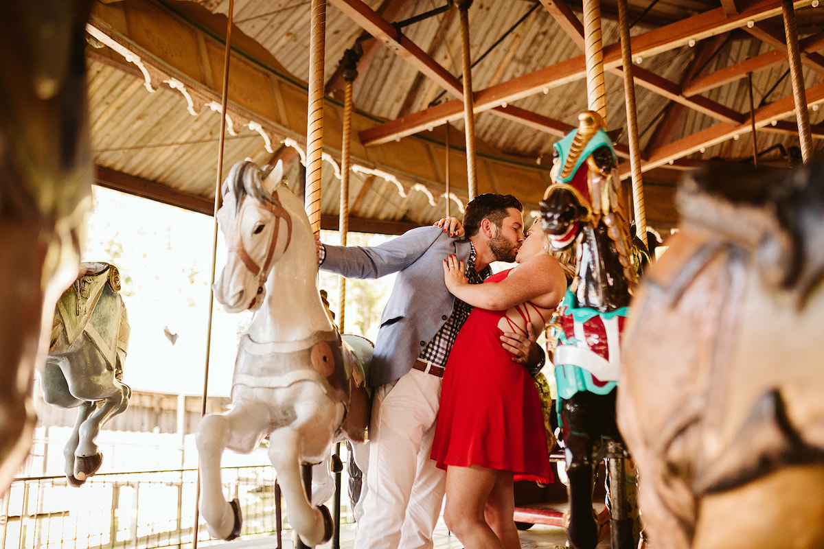 Man holds onto carousel horse and leans into his fiancé to kiss her