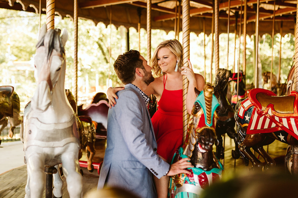 Woman sits on carousel horse and gazes at man standing next to her for their fun engagement photos at Lake Winnie