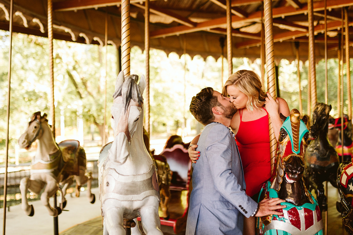 Woman in red dress sits on carousel horse and leans over to kiss man in blue blazer standing next to her