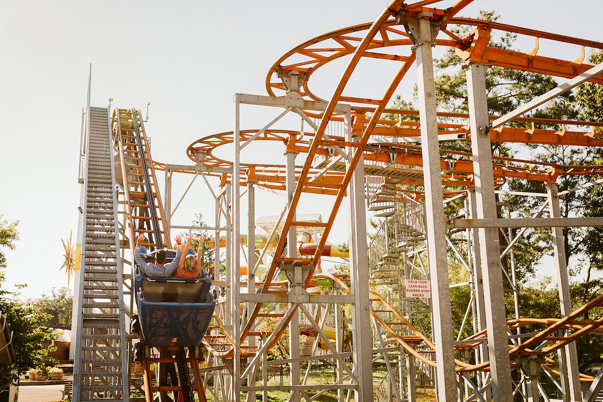 Man and woman ride in roller coaster with their hands raised.