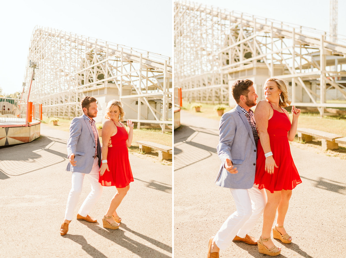 Man in white pants and blue sport coat dances with woman in red dress below rollercoaster for fun engagement photos