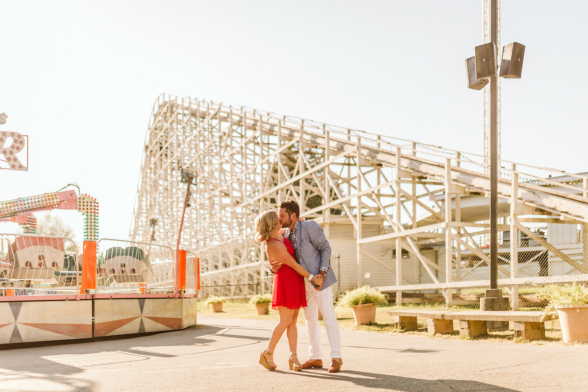 Man in white pants and blue sport coat kisses woman in red dress below rollercoaster framing for fun engagement photos
