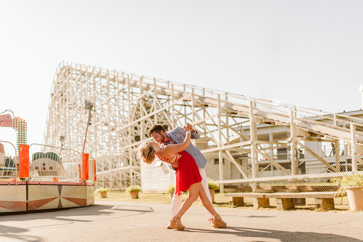 Man in white pants and blue sport coat dances with woman in red dress below rollercoaster for fun engagement photos