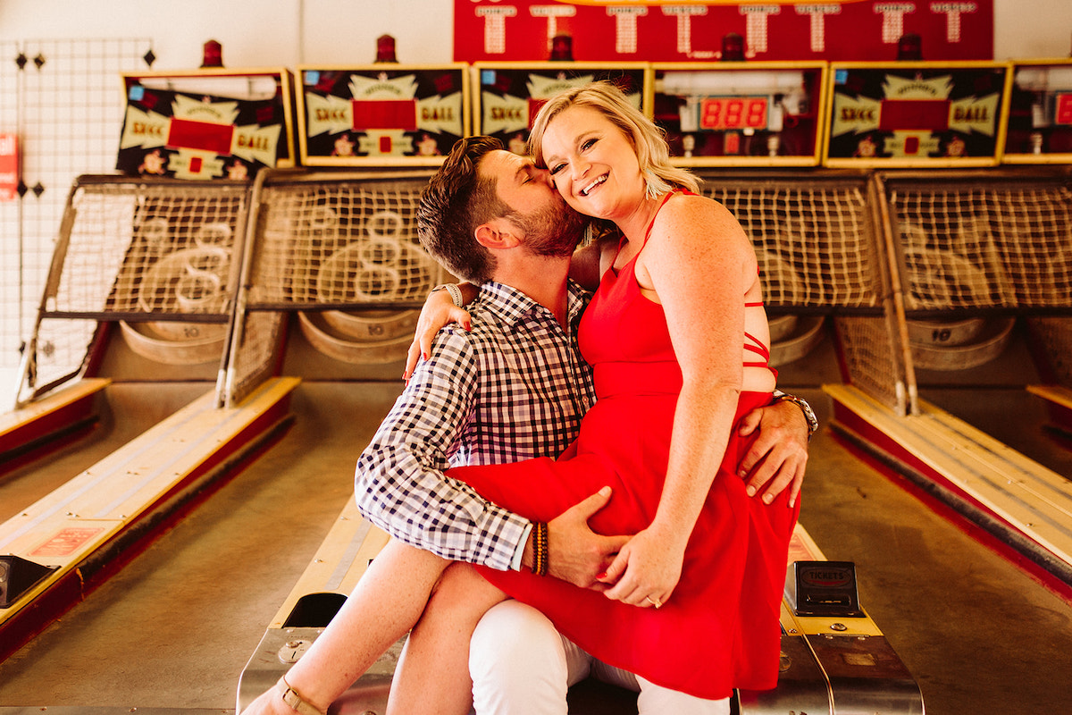 Man sits with woman on his lap at the end of Skee-ball game at the Lake Winnie arcade for their unique engagement photos