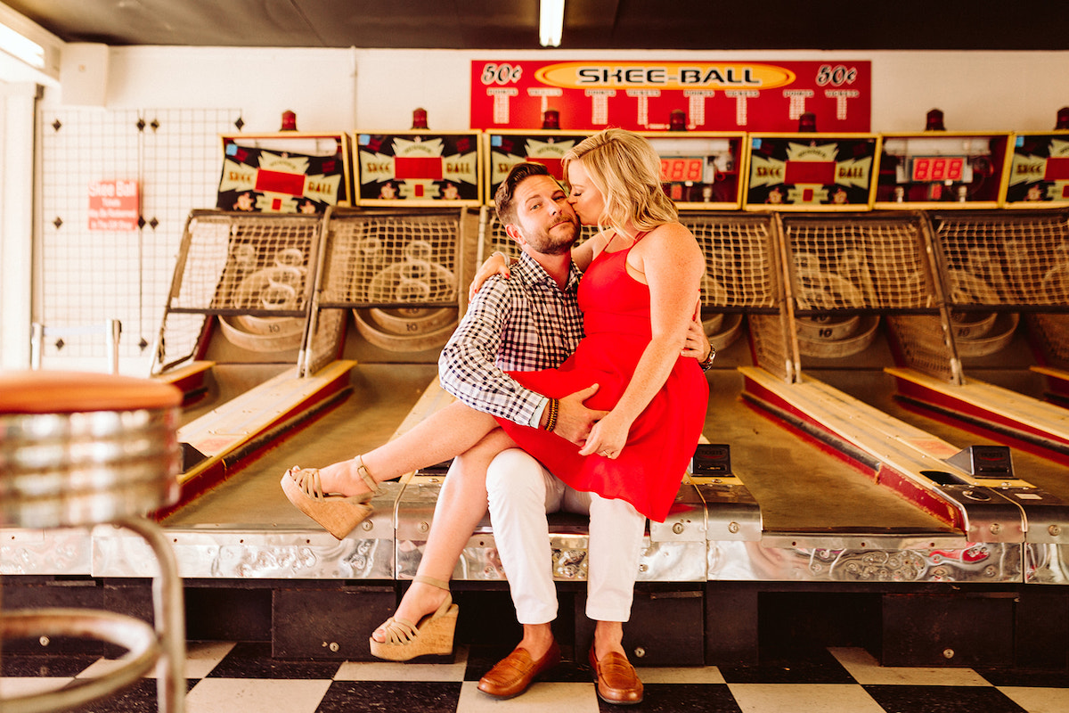 Woman kisses man's cheek as she sits on his lap on the Skee-ball arcade game for their cute engagement photos in Chattanooga