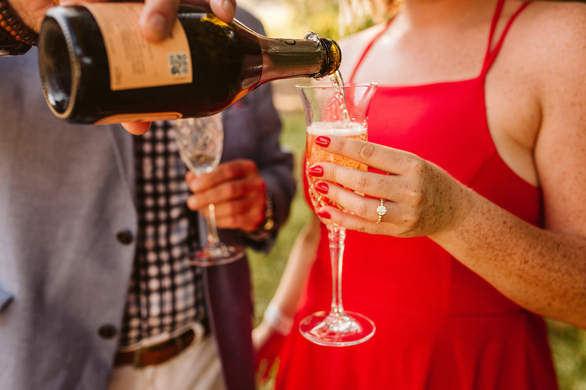 Woman holds crystal glass, her engagement ring displayed, while man pours champagne into it