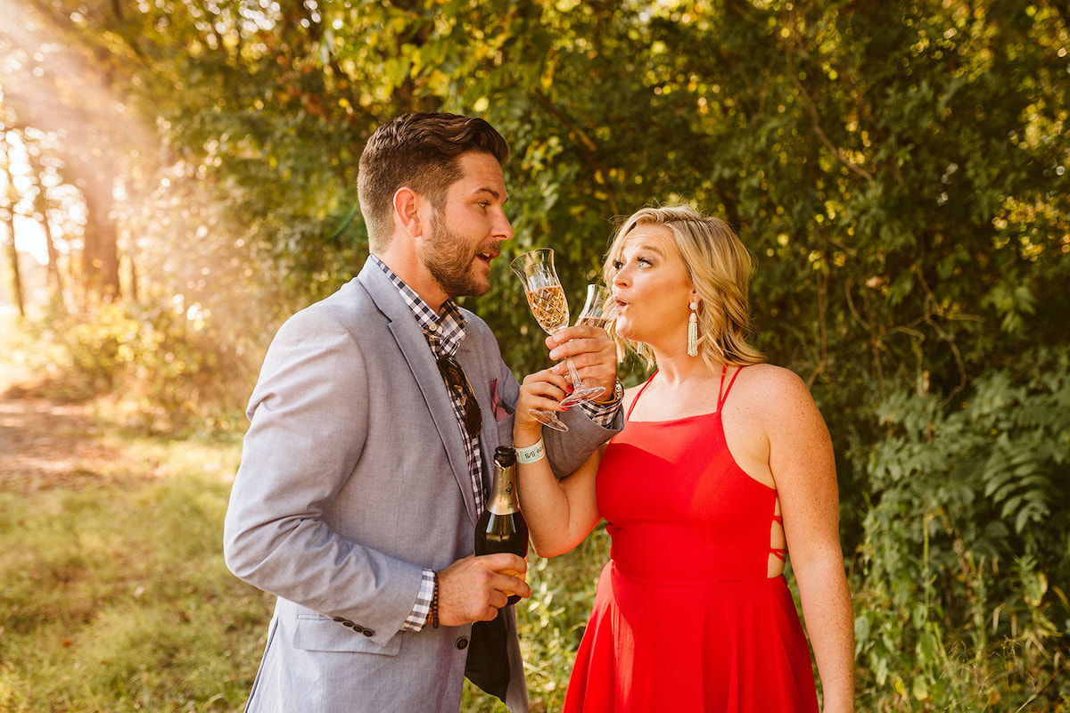 Man in light blue sport coat and woman in red dress link arms to drink champagne from crystal glasses