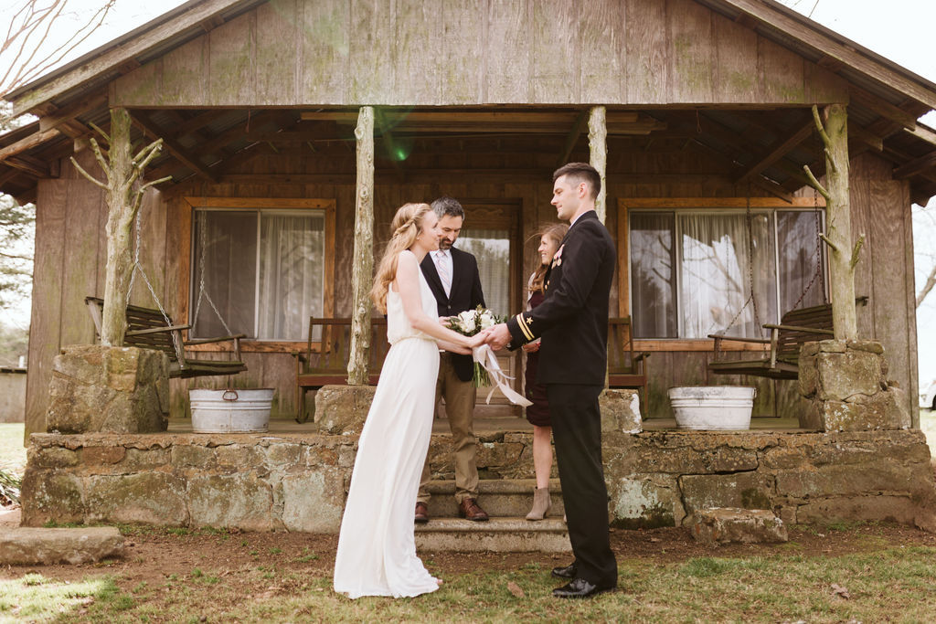 Bride and groom stand facing each other holding hands and exchanging vows in front of antique family cabin front porch