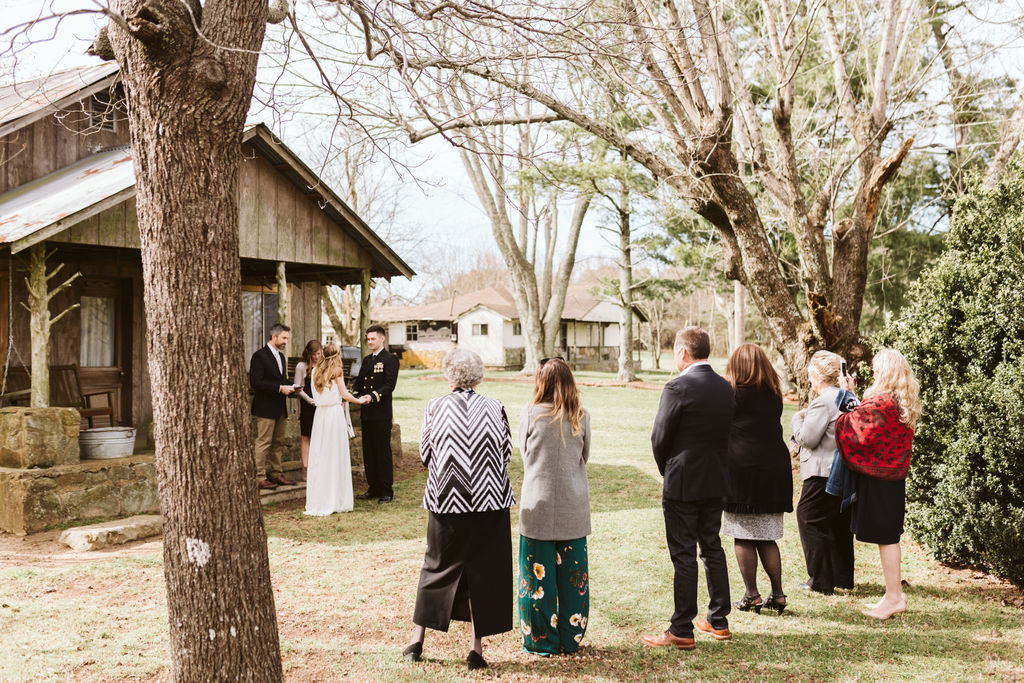 Bride and groom facing each other holding hands in front of antique family cabin porch while family members stand nearby