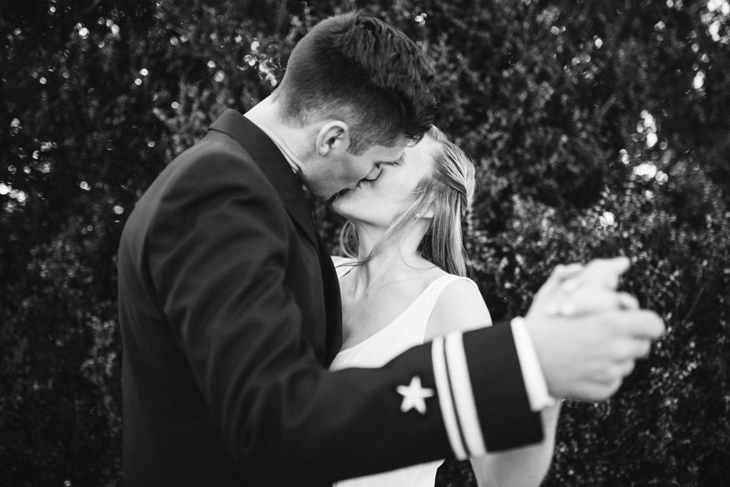 Bride and groom share a dance in front of greenery, his head bowed over to kiss her