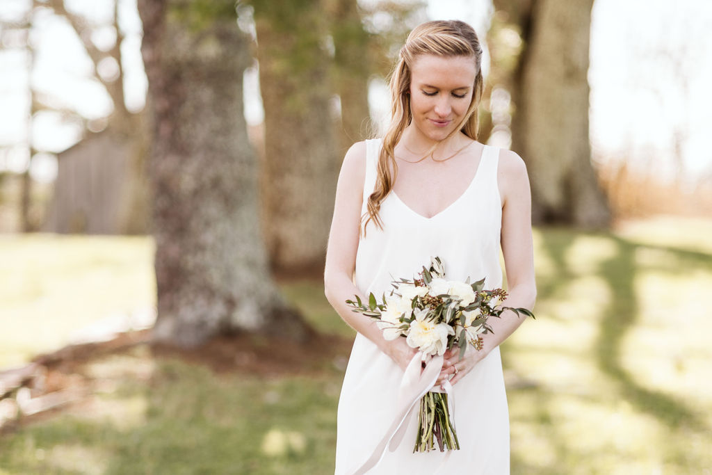Bride stands holding her bouquet in front of her smiling down at it, trees in the background