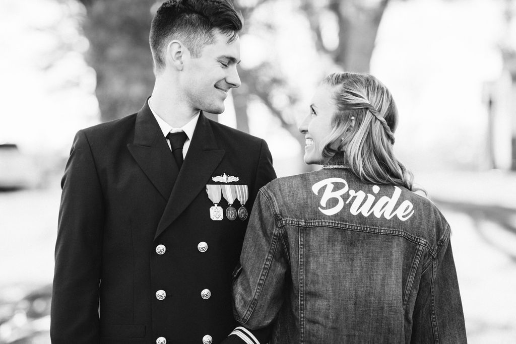 Bride and groom stand arm in arm. He wears a military dress uniform, and she wears a customized bride denim jacket.