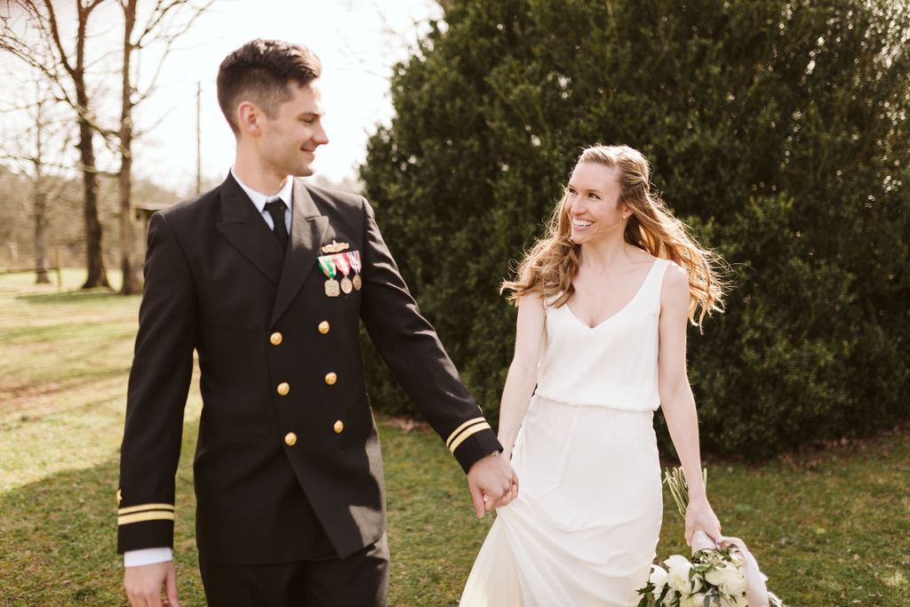 Groom in military dress uniform holds his bride's hand as they smile at each other