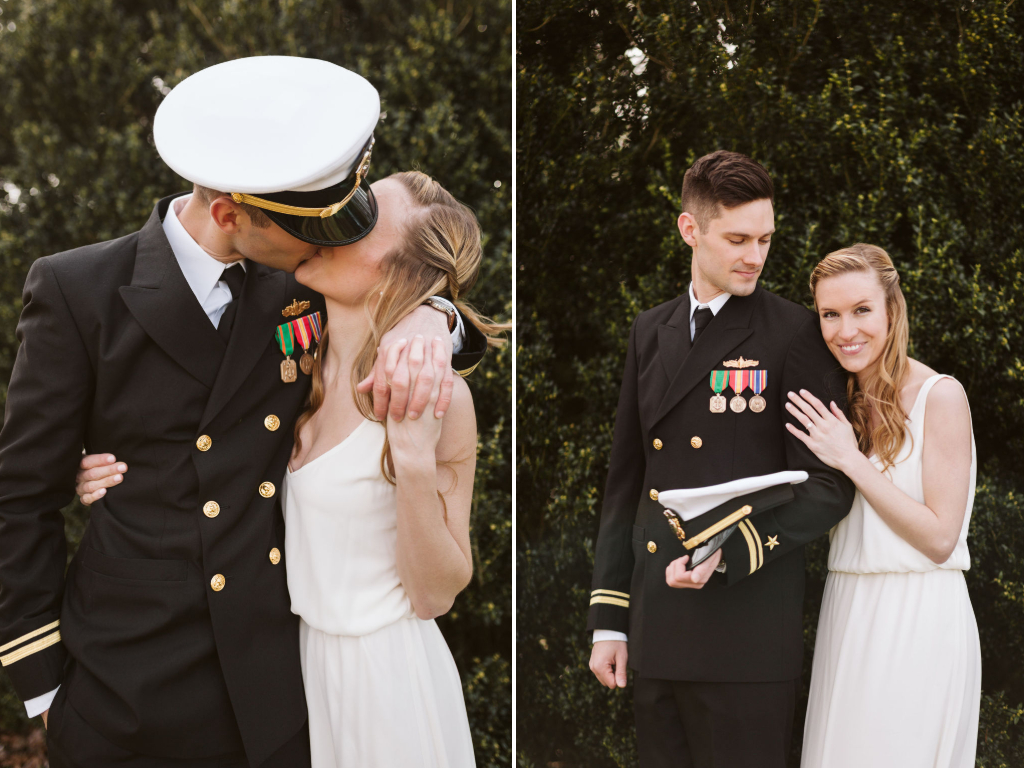 Bride wearing simple white dress and groom wearing military dress uniform and hat cuddle and kiss in front of greenery.