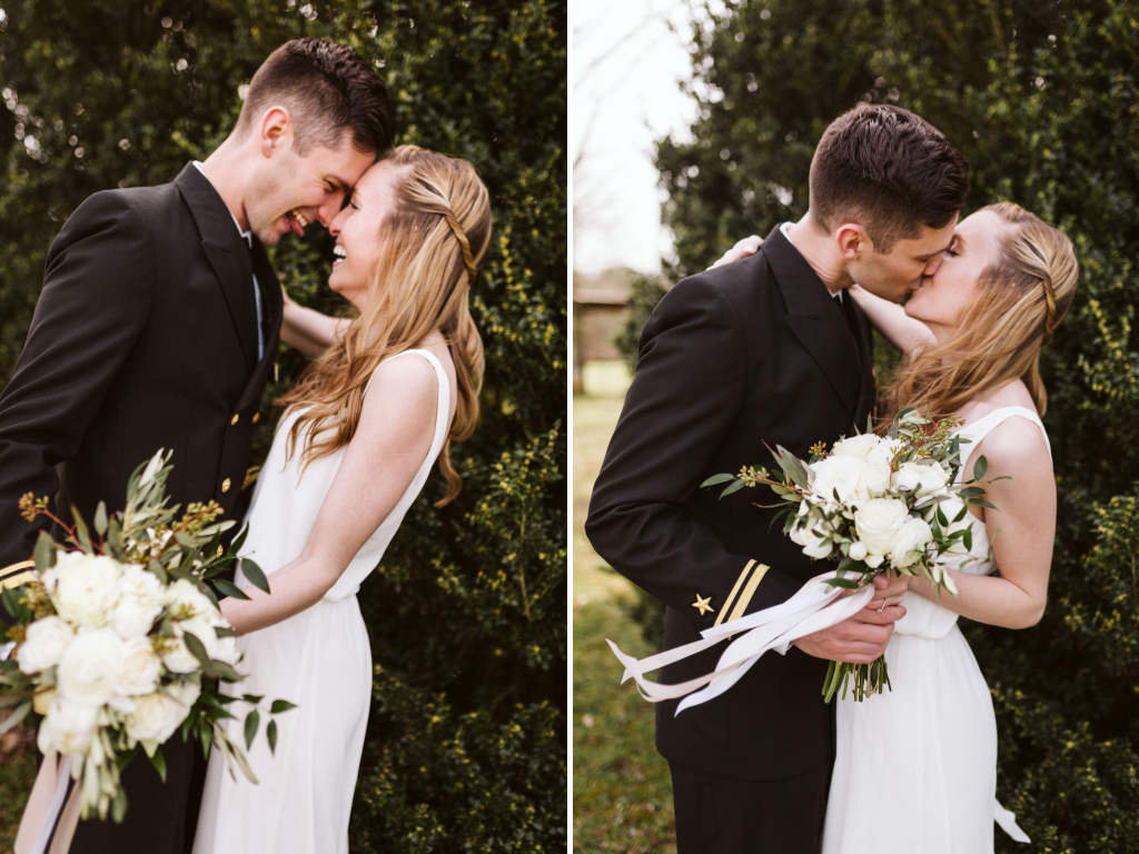 Bride wearing simple white dress and groom wearing military dress uniform and hat cuddle and kiss in front of greenery.