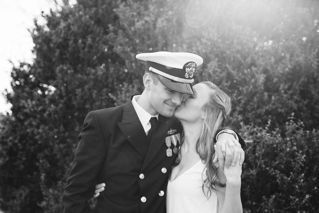 Groom wearing military dress uniform and hat leans down so his bride can reach his cheek for a kiss.