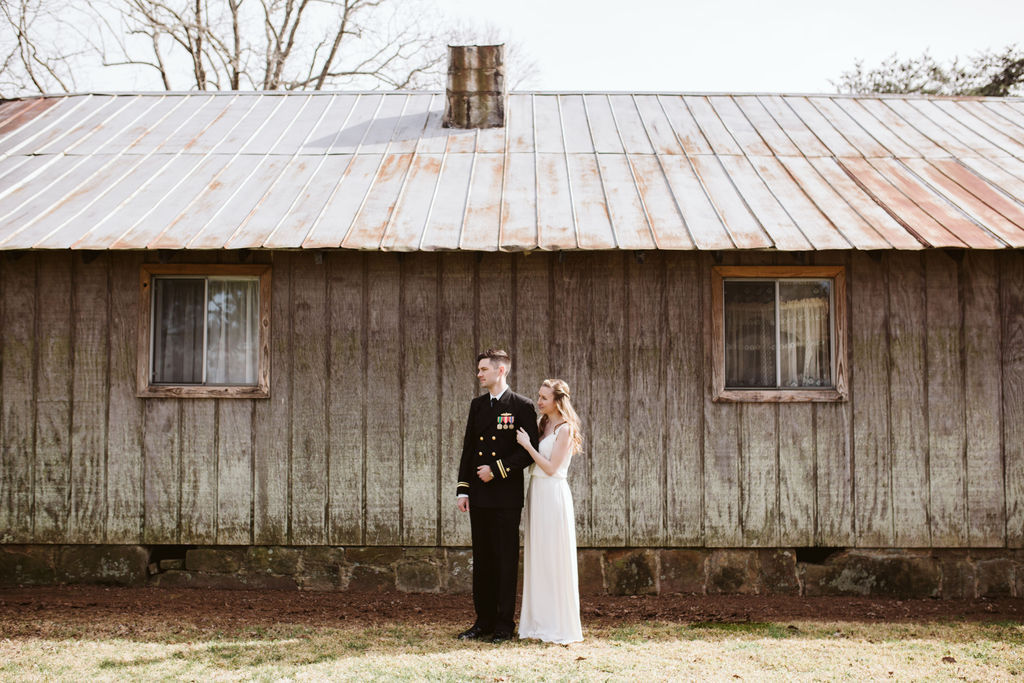 Bride and groom cuddle arm-in-arm in front of antique family cabin after their intimate elopement