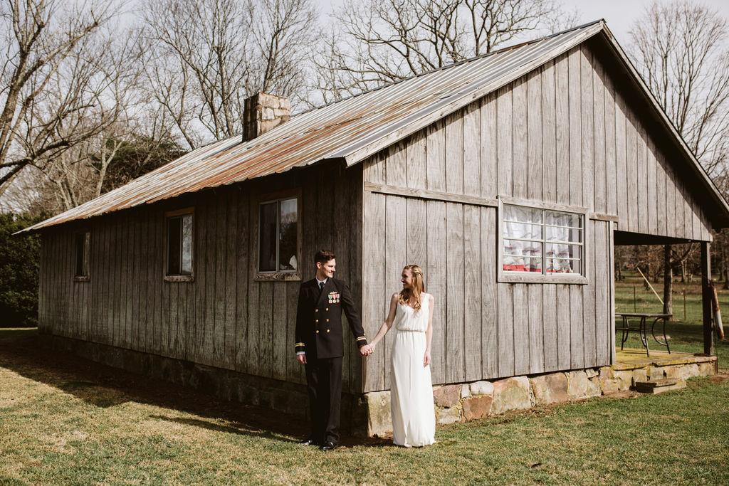 Bride and groom hold hands and look at each other in front of antique family cabin after their intimate elopement