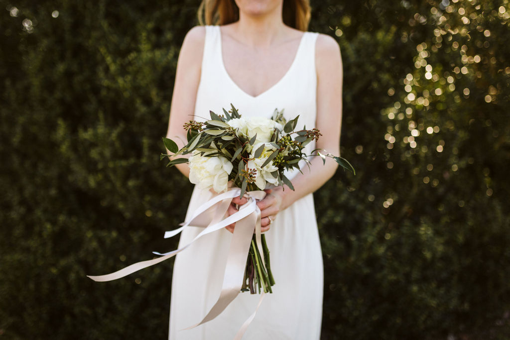 Bride holds her bouquet in front of her, white flowers and greenery with champagne colored ribbons blowing in the breeze.