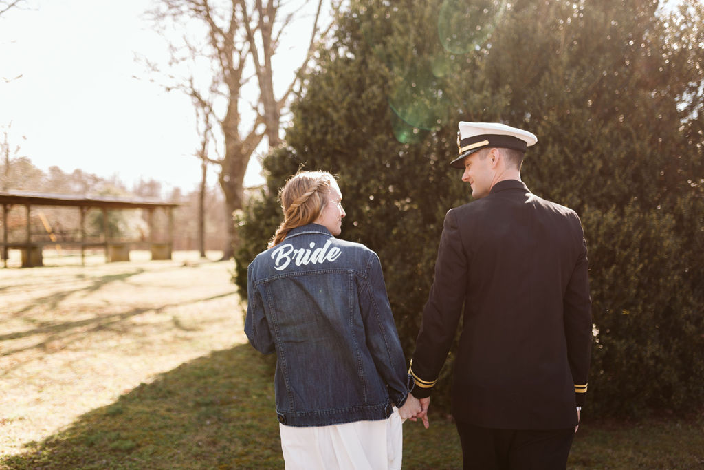 Bride and groom hold hands, walking away. He wears a military dress uniform, and she wears a customized bride denim jacket