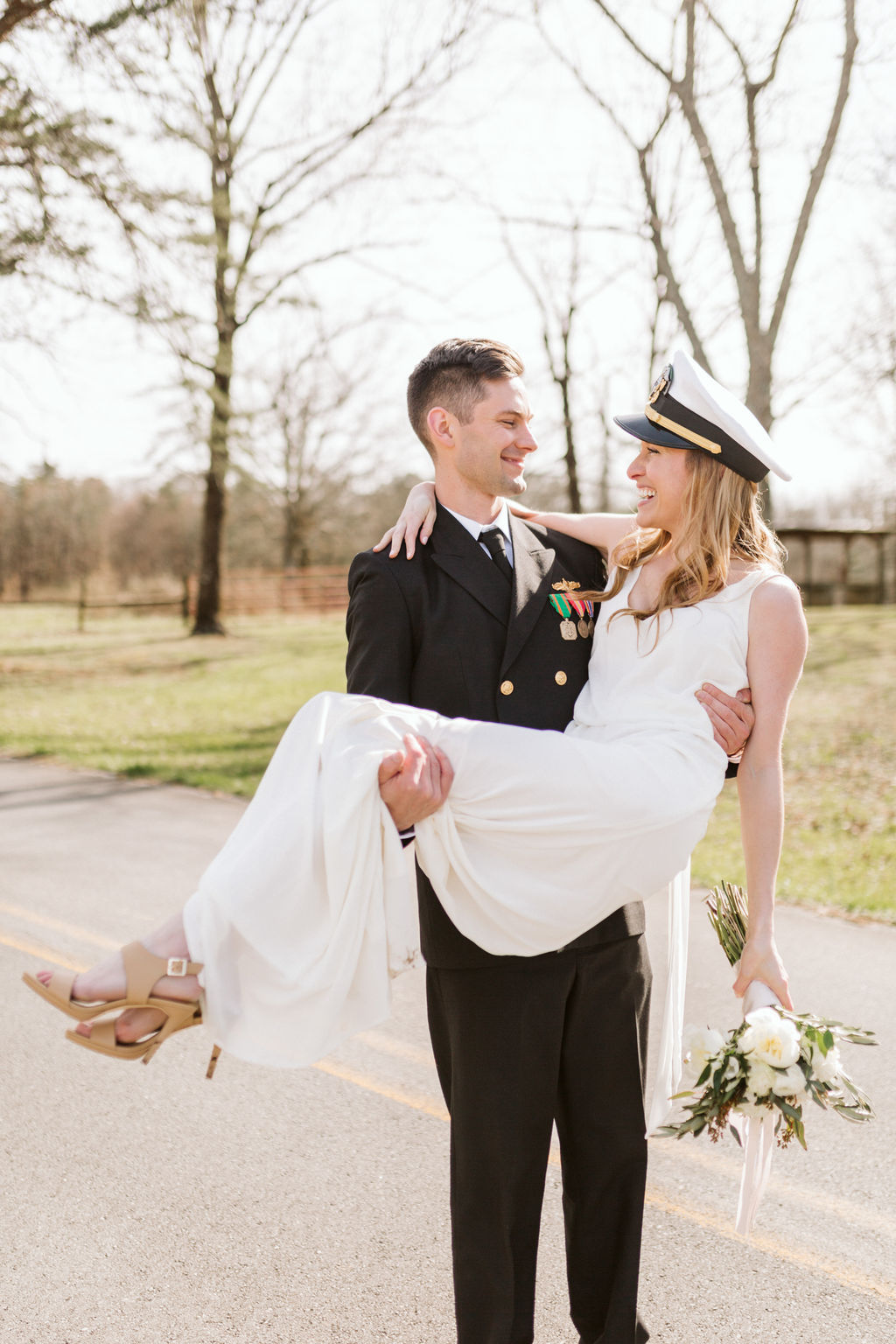 Groom in military dress uniform holds his bride in his arms. She wears his military cap and holds a bouquet at her side.