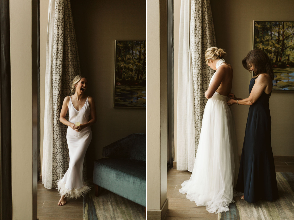 Woman in white feather-hemmed slip laughing in front of floor to ceiling window and mother fastening waist on wedding dress